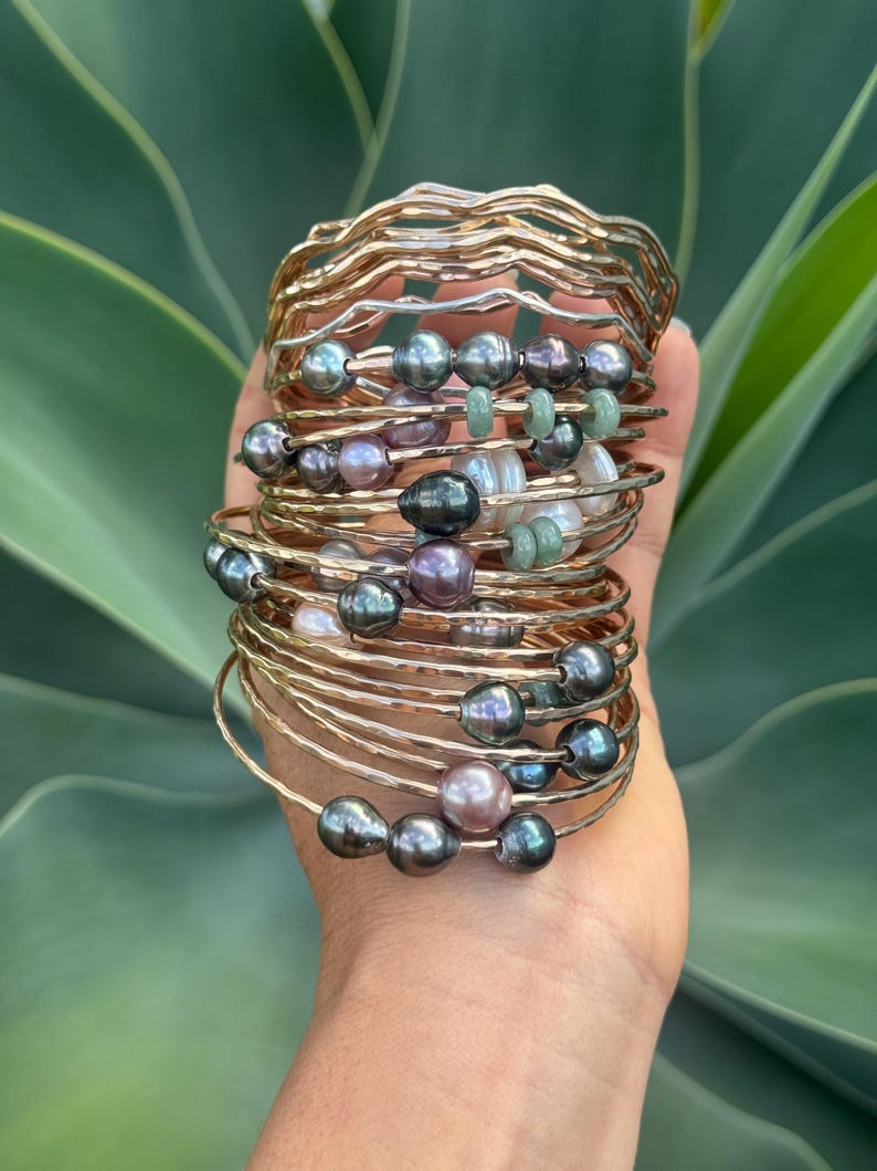 May include: A stack of gold-toned bangles, some with a hammered texture, adorned with dark gray, lavender, and white pearls, and small green beads. The bracelets are stacked on top of each other, creating a layered effect.
