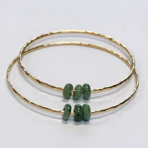 May include: Two gold-colored bangles with a hammered texture. Each bangle features three green, oval-shaped beads. The jewelry is set against a white background, highlighting the contrast between the gold and green.