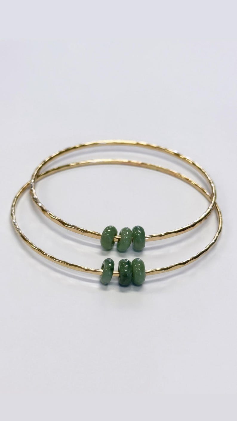 May include: Two gold-colored bangles with a hammered texture. Each bangle features three green, oval-shaped beads. The jewelry is set against a white background, highlighting the contrast between the gold and green.