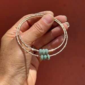 May include: Two gold-colored bangles with a hammered texture are held in a hand. The bangles are adorned with six small, light green beads. The background is a solid, reddish-brown color.