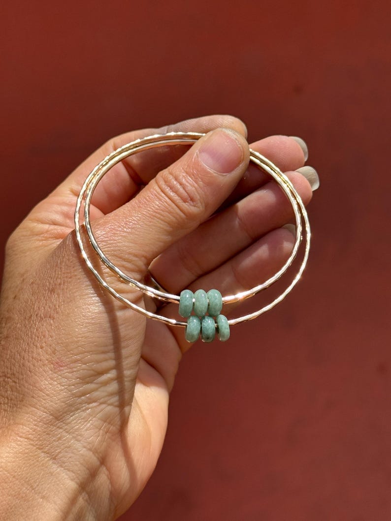 May include: Two gold-colored bangles with a hammered texture are held in a hand. The bangles are adorned with six small, light green beads. The background is a solid, reddish-brown color.