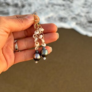 May include: Gold-toned dangle earrings featuring a combination of small white shells, gold chain links, and dark gray pearls. Each earring has a small, clear gemstone accent. The earrings are held up against a blurred ocean background.