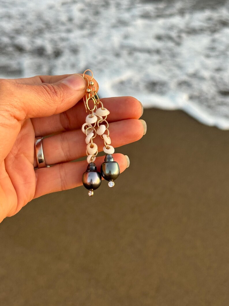 May include: Gold-toned dangle earrings featuring a combination of small white shells, gold chain links, and dark gray pearls. Each earring has a small, clear gemstone accent. The earrings are held up against a blurred ocean background.