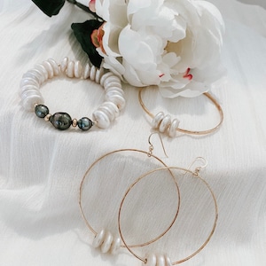 K&ouml;nnte beinhalten: Ein Schmuckset bestehend aus einem elastischen Armband mit wei&szlig;en und dunkelgrauen M&uuml;nzperlen, einem d&uuml;nnen Goldarmreif mit drei wei&szlig;en M&uuml;nzperlen und einem Paar gro&szlig;er Goldcreolen, die jeweils mit drei wei&szlig;en M&uuml;nzperlen verziert sind.