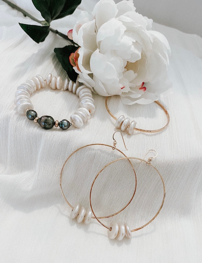 K&ouml;nnte beinhalten: Ein Schmuckset bestehend aus einem elastischen Armband mit wei&szlig;en und dunkelgrauen M&uuml;nzperlen, einem d&uuml;nnen Goldarmreif mit drei wei&szlig;en M&uuml;nzperlen und einem Paar gro&szlig;er Goldcreolen, die jeweils mit drei wei&szlig;en M&uuml;nzperlen verziert sind.