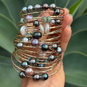 May include: A collection of gold-toned bangles, each adorned with a variety of beads. The beads include dark gray, light pink, and white pearls, as well as small green stones. The bangles are stacked together, creating a layered effect.