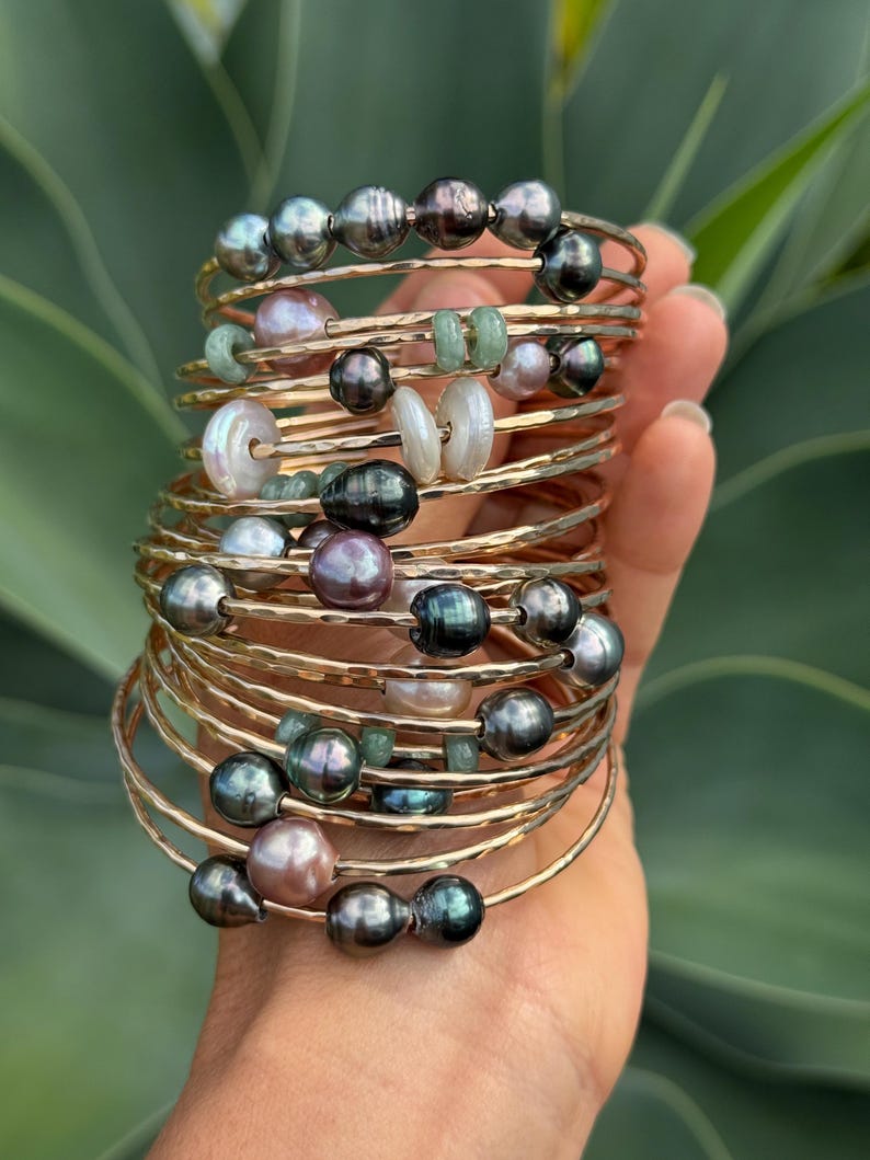 May include: A collection of gold-toned bangles, each adorned with a variety of beads. The beads include dark gray, light pink, and white pearls, as well as small green stones. The bangles are stacked together, creating a layered effect.