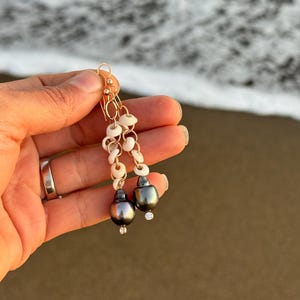 May include: Gold dangle earrings featuring dark gray pearls, small white shell beads, and tiny clear crystals. The earrings are held up against a blurred background of the ocean and sand.