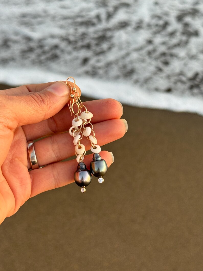May include: Gold dangle earrings featuring dark gray pearls, small white shell beads, and tiny clear crystals. The earrings are held up against a blurred background of the ocean and sand.