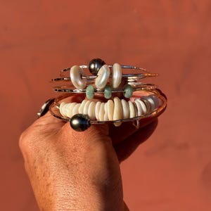 K&ouml;nnte beinhalten: Ein Stapel aus vier d&uuml;nnen Silberarmreifen mit verschiedenen Perlen. Die Armb&auml;nder sind mit dunkelgrauen Perlen, wei&szlig;en m&uuml;nzf&ouml;rmigen Perlen und kleinen gr&uuml;nen Steinabstandshaltern verziert. Die Armreifen haben eine geh&auml;mmerte Textur.