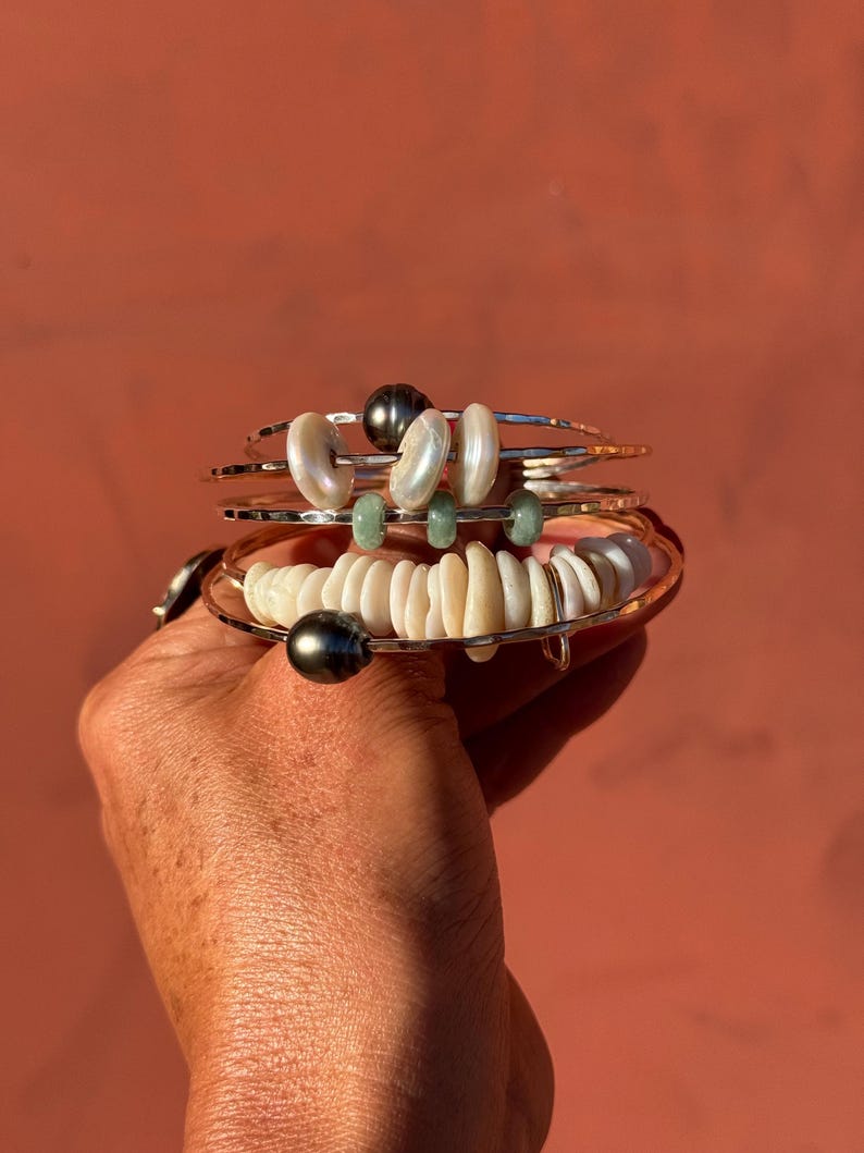 K&ouml;nnte beinhalten: Ein Stapel aus vier d&uuml;nnen Silberarmreifen mit verschiedenen Perlen. Die Armb&auml;nder sind mit dunkelgrauen Perlen, wei&szlig;en m&uuml;nzf&ouml;rmigen Perlen und kleinen gr&uuml;nen Steinabstandshaltern verziert. Die Armreifen haben eine geh&auml;mmerte Textur.
