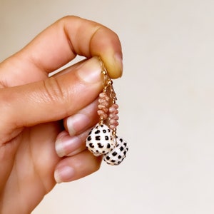 May include: Dangle earrings featuring small, white, spotted sea shells suspended from gold-toned wire. Above each shell is a cluster of tiny, textured, light pink, cylindrical beads. The earrings are attached to gold-toned metal ear hooks.