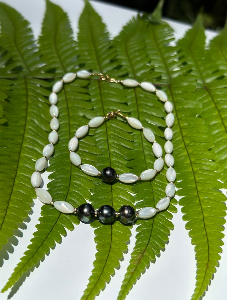 May include: Two necklaces with white oval beads and black pearls. The necklaces are on a green fern leaf background.