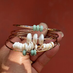 May include: A set of gold-toned bangles with decorative elements. Some bangles feature light green beads, while others have white shell-like pieces. The bangles are held against a reddish-brown background.