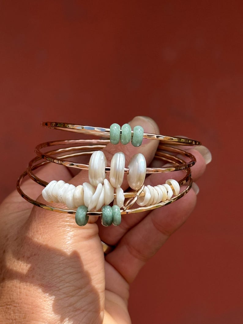 May include: A set of gold-toned bangles with decorative elements. Some bangles feature light green beads, while others have white shell-like pieces. The bangles are held against a reddish-brown background.
