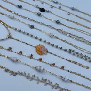May include: A collection of delicate gold necklaces with various natural elements. Some necklaces feature pearls, seashells, and small gemstones. The necklaces are arranged on a white surface, showcasing their intricate designs and textures.