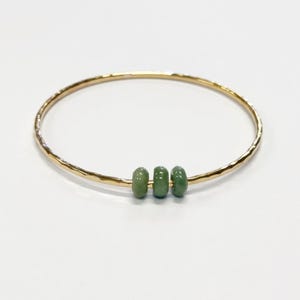 May include: A gold-colored bangle bracelet with a hammered texture. The bracelet features three small, oval-shaped, green beads. The jewelry is displayed on a white background.