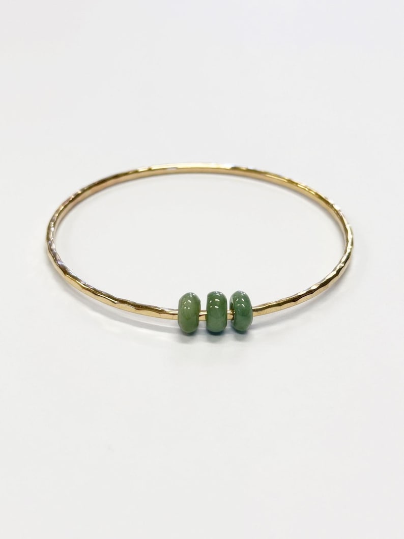 May include: A gold-colored bangle bracelet with a hammered texture. The bracelet features three small, oval-shaped, green beads. The jewelry is displayed on a white background.