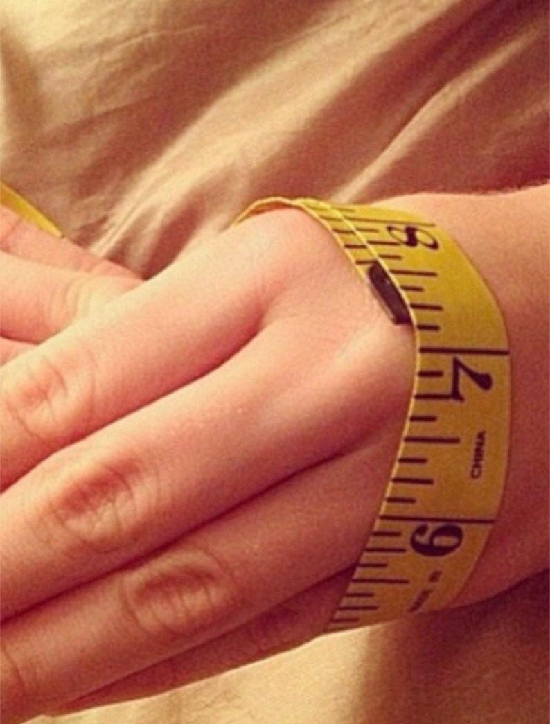 May include: A yellow measuring tape wrapped around a wrist, showing measurements in inches. The tape is marked with black numbers and lines, indicating a measurement of approximately 6.5 inches.