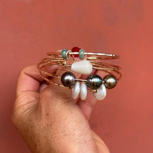 K&ouml;nnte beinhalten: Ein mehrreihiges Armband aus geh&auml;mmertem, goldfarbenem Metall mit verschiedenen Anh&auml;ngern, darunter zwei kleine gr&uuml;ne Perlen, eine wei&szlig;e tropfenf&ouml;rmige Perle, drei dunkelgraue runde Perlen und drei kleine, flache, scheibenf&ouml;rmige wei&szlig;e Perlen.