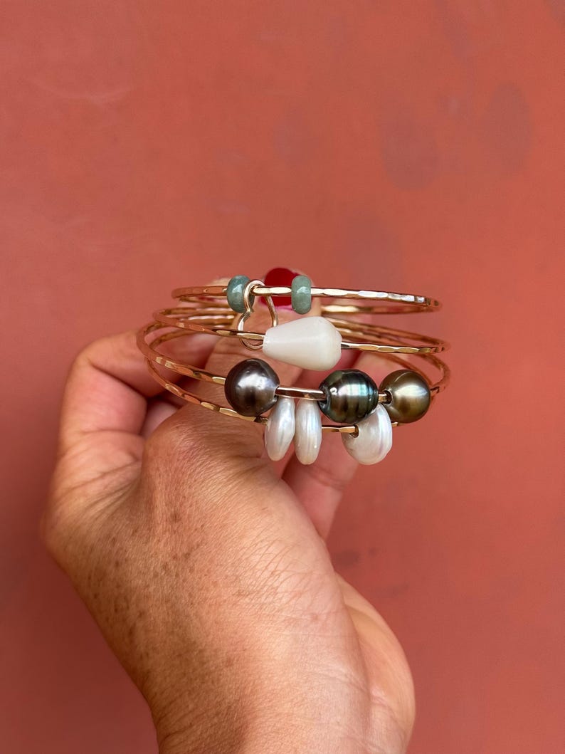 K&ouml;nnte beinhalten: Ein mehrreihiges Armband aus geh&auml;mmertem, goldfarbenem Metall mit verschiedenen Anh&auml;ngern, darunter zwei kleine gr&uuml;ne Perlen, eine wei&szlig;e tropfenf&ouml;rmige Perle, drei dunkelgraue runde Perlen und drei kleine, flache, scheibenf&ouml;rmige wei&szlig;e Perlen.