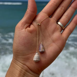 May include: A gold-colored lariat necklace featuring two seashell pendants. One pendant is white and tan, the other is purple and tan. The necklace is held in a hand with a silver ring, with the ocean in the background.