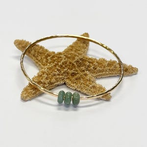 May include: A gold-colored bangle bracelet with three small, round, light green beads. The bracelet is resting on a tan starfish against a white background. The bangle has a hammered texture.