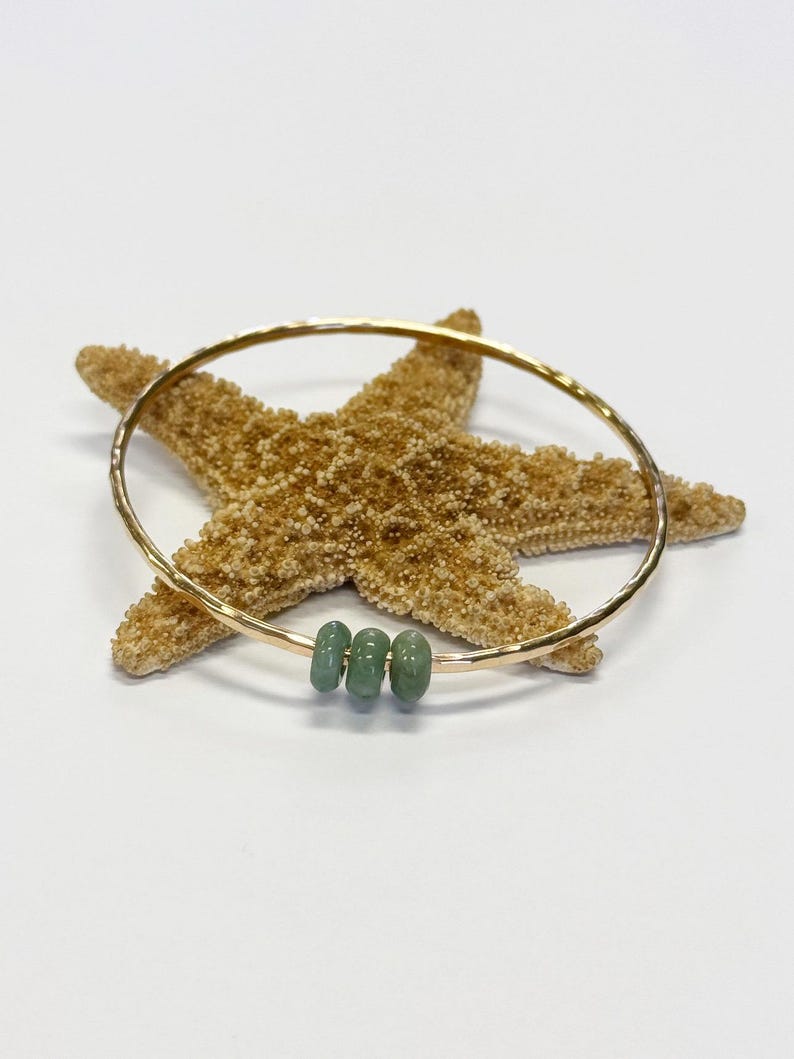May include: A gold-colored bangle bracelet with three small, round, light green beads. The bracelet is resting on a tan starfish against a white background. The bangle has a hammered texture.