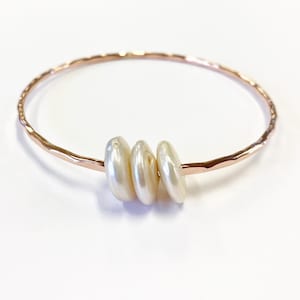 K&ouml;nnte beinhalten: Ein Armreif aus Ros&eacute;gold mit einer geh&auml;mmerten, strukturierten Metalloberfl&auml;che. Drei glatte, cremewei&szlig;e, m&uuml;nzf&ouml;rmige Perlen sind auf den kreisf&ouml;rmigen Reifen gef&auml;delt und befinden sich zusammen in der Mitte des Metalldrahtrahmens.