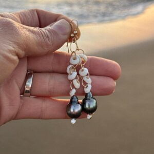 May include: Gold dangle earrings featuring dark gray teardrop-shaped pearls with small clear crystals at the base. The earrings also have white shell accents and gold-colored hooks.