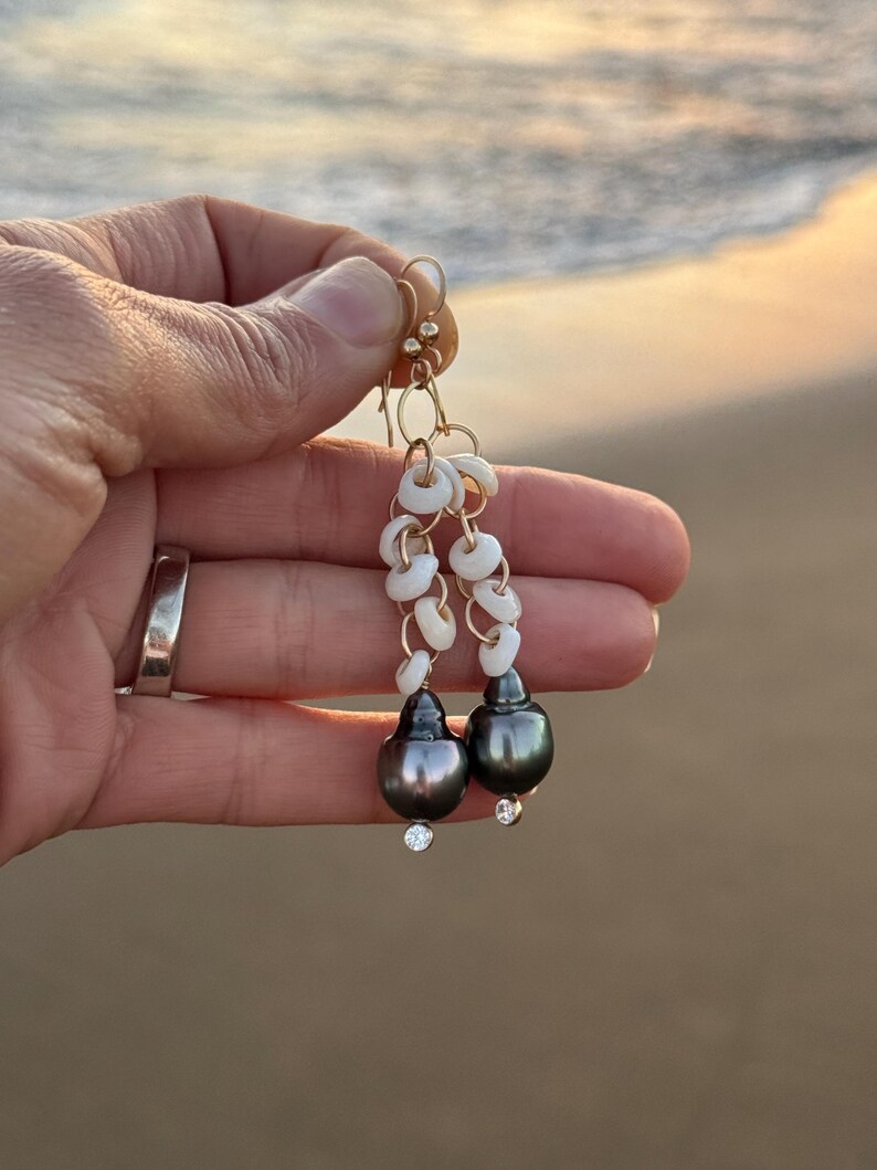 May include: Gold dangle earrings featuring dark gray teardrop-shaped pearls with small clear crystals at the base. The earrings also have white shell accents and gold-colored hooks.