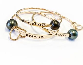 May include: Three gold bangles with black pearl accents. The bangles are engraved with the names "Aina Rose", "Moaniani", and "Waianuhe"