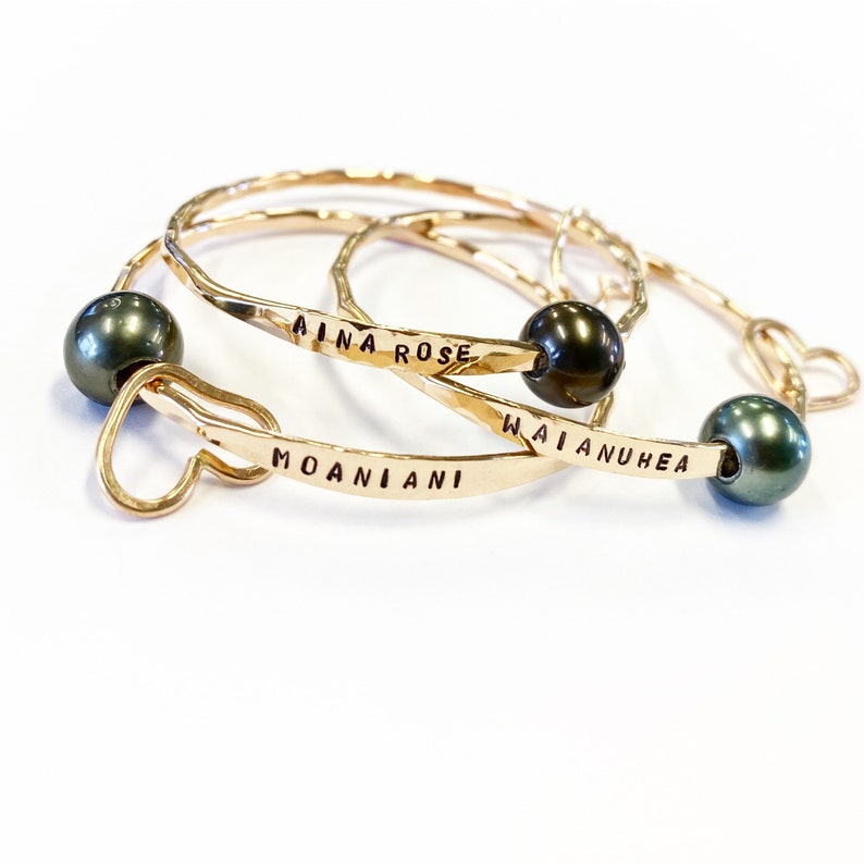Puede incluir: Tres brazaletes de oro con detalles de perlas negras. Los brazaletes est&aacute;n grabados con los nombres "Aina Rose", "Moaniani" y "Waianuhe"