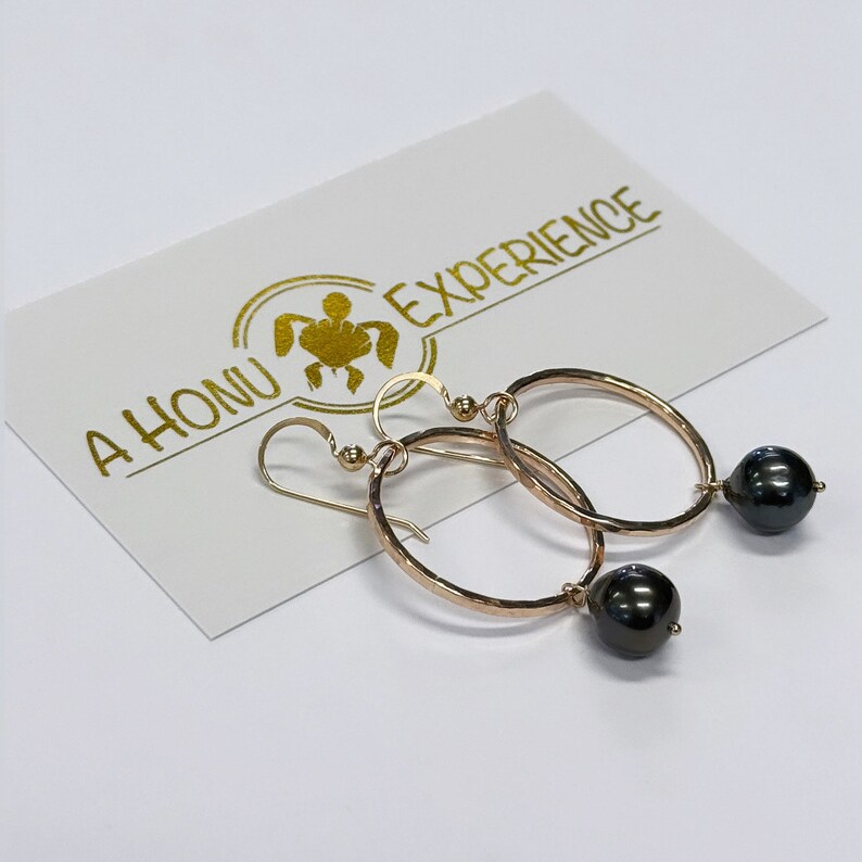 May include: A pair of gold hoop earrings with black pearl accents. The earrings are hammered and have a textured finish. The earrings are hanging from a white card with the text "A HONU EXPERIENCE".
