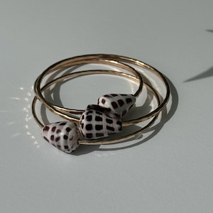 May include: A set of three gold-colored bangles, each adorned with a seashell bead. The seashells are white with a brown checkered pattern. The bangles are stacked on a white surface, with shadows cast across the scene.