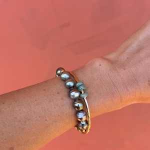 May include: Two bracelets on a wrist. One is a gold bangle with a strand of gray pearls and green beads. The other is a gold bangle. The background is a solid coral color.