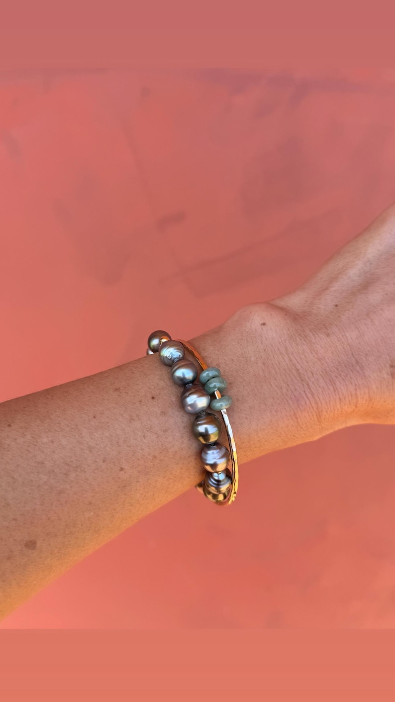 May include: Two bracelets on a wrist. One is a gold bangle with a strand of gray pearls and green beads. The other is a gold bangle. The background is a solid coral color.