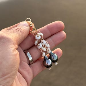 May include: Gold-toned dangle earrings featuring white shell accents and dark gray pearls. The earrings are held in a hand, showcasing the delicate design and craftsmanship. The pearls have a subtle luster, adding elegance to the jewelry.