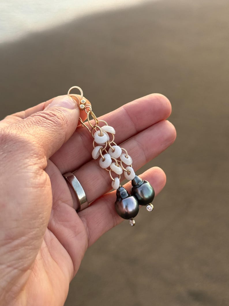 May include: Gold-toned dangle earrings featuring white shell accents and dark gray pearls. The earrings are held in a hand, showcasing the delicate design and craftsmanship. The pearls have a subtle luster, adding elegance to the jewelry.