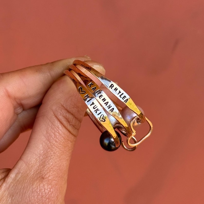 Puede incluir: Tres pulseras de brazalete doradas con los nombres grabados: "Rhylea", "Kamehana" y "Miyuki" con un s&iacute;mbolo de coraz&oacute;n en cada una. Las pulseras est&aacute;n apiladas una encima de la otra y tienen un dije de perla negra.