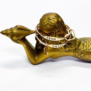 Puede incluir: Dos pulseras de brazalete personalizadas doradas con los nombres "Bryce" y "Valley" grabados en ellas. Las pulseras se muestran en una estatua de sirena dorada.