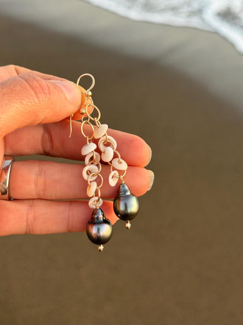 May include: Gold dangle earrings featuring dark gray pearls and small white shell accents. The earrings are suspended from gold chains and hooks, creating a layered effect. The background is a blurred view of a beach.