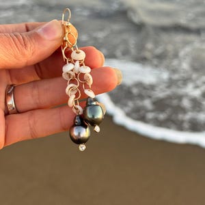 May include: Gold-tone dangle earrings featuring dark gray pearls with small clear crystals. The earrings also include small white shell accents and a gold-tone circular design. The background shows a blurred ocean scene.