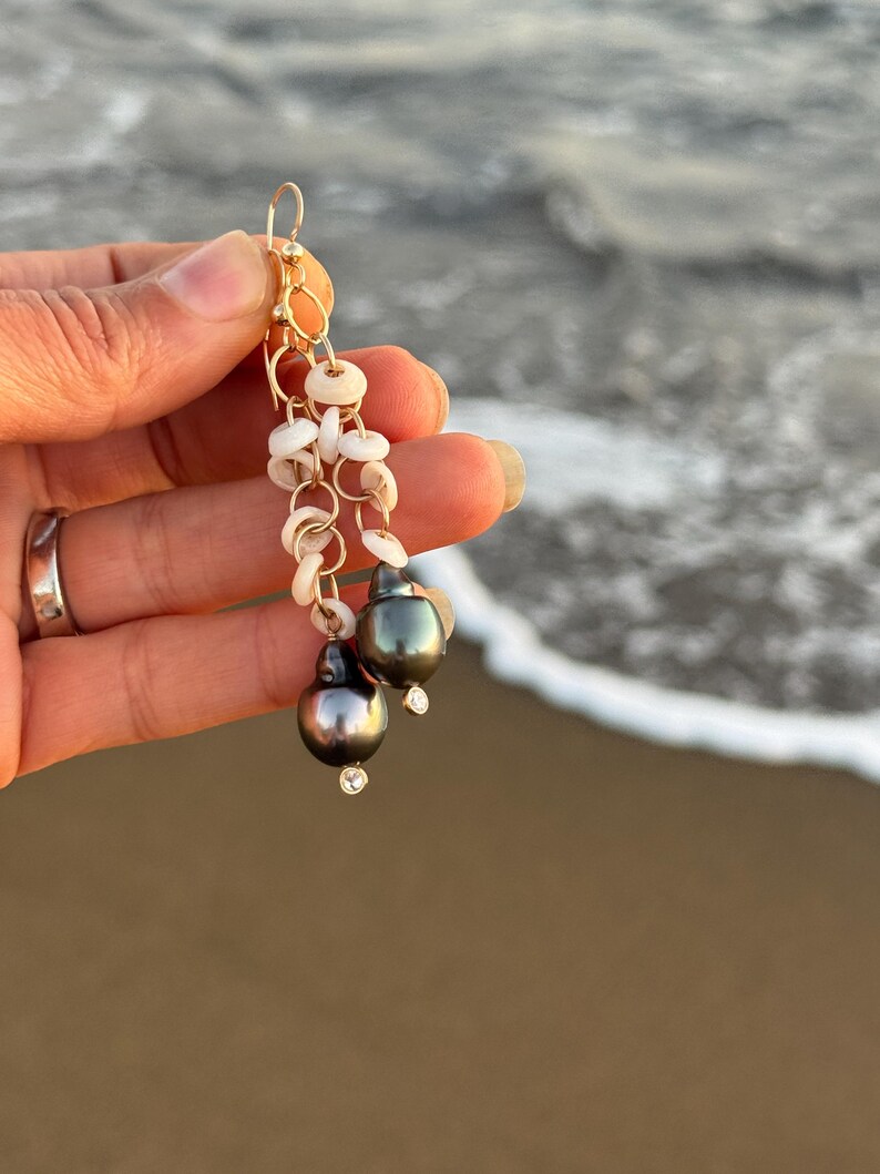 May include: Gold-tone dangle earrings featuring dark gray pearls with small clear crystals. The earrings also include small white shell accents and a gold-tone circular design. The background shows a blurred ocean scene.