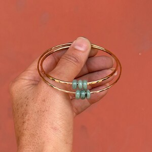 May include: Three gold-colored bangles with a hammered texture are held against a reddish-orange background. Each bangle has a set of three light green beads. The bangles are simple and elegant.
