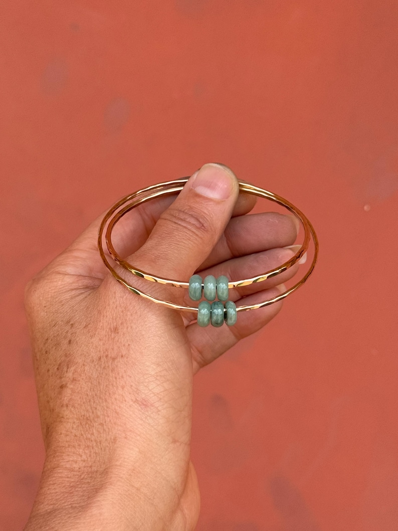 May include: Three gold-colored bangles with a hammered texture are held against a reddish-orange background. Each bangle has a set of three light green beads. The bangles are simple and elegant.