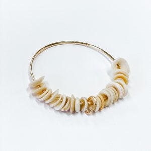 May include: A thin gold-toned metal hoop bracelet adorned with a series of small, off-white, circular shell beads. The beads are strung along the lower portion of the hoop, interspersed with a single small gold-toned heart-shaped metal charm.