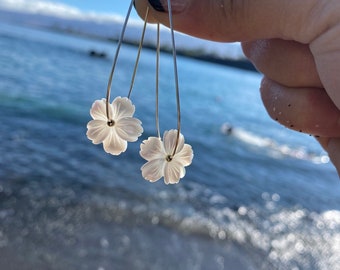 May include: A pair of delicate, white flower earrings with silver-tone wire hooks. The earrings are held up against a backdrop of a blue ocean and sky. The flower petals have a subtle, pearlescent sheen.