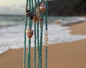May include: A multi-strand necklace featuring turquoise-colored seed beads interspersed with small gold-toned spacer beads. The strands are accented with various natural shells, including a striped spiral shell, a smooth pink pearl, and white petal-shaped shells.