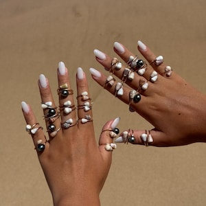 May include: A collection of thin metal rings worn on multiple fingers, each featuring small white or tan seashells and dark grey or black spherical beads. The rings are arranged in stacks and single bands across both hands, showcasing a variety of natural shell shapes and smooth, dark, round accents set on simple metallic bands.